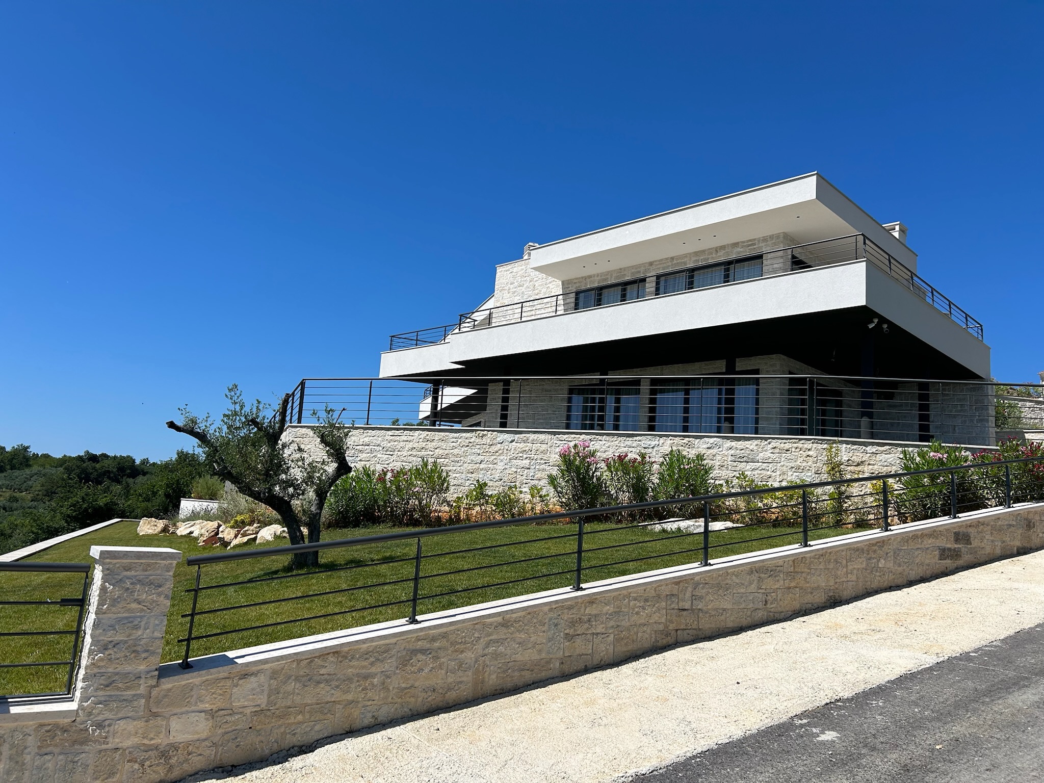 Moderna dvokatnica s kamenim i bijelim pročeljem, velikim balkonom i uređenim dvorištem, uz vedro plavo nebo. Elko - Instalacija Pametnih Kuća i Smart Home Sistema