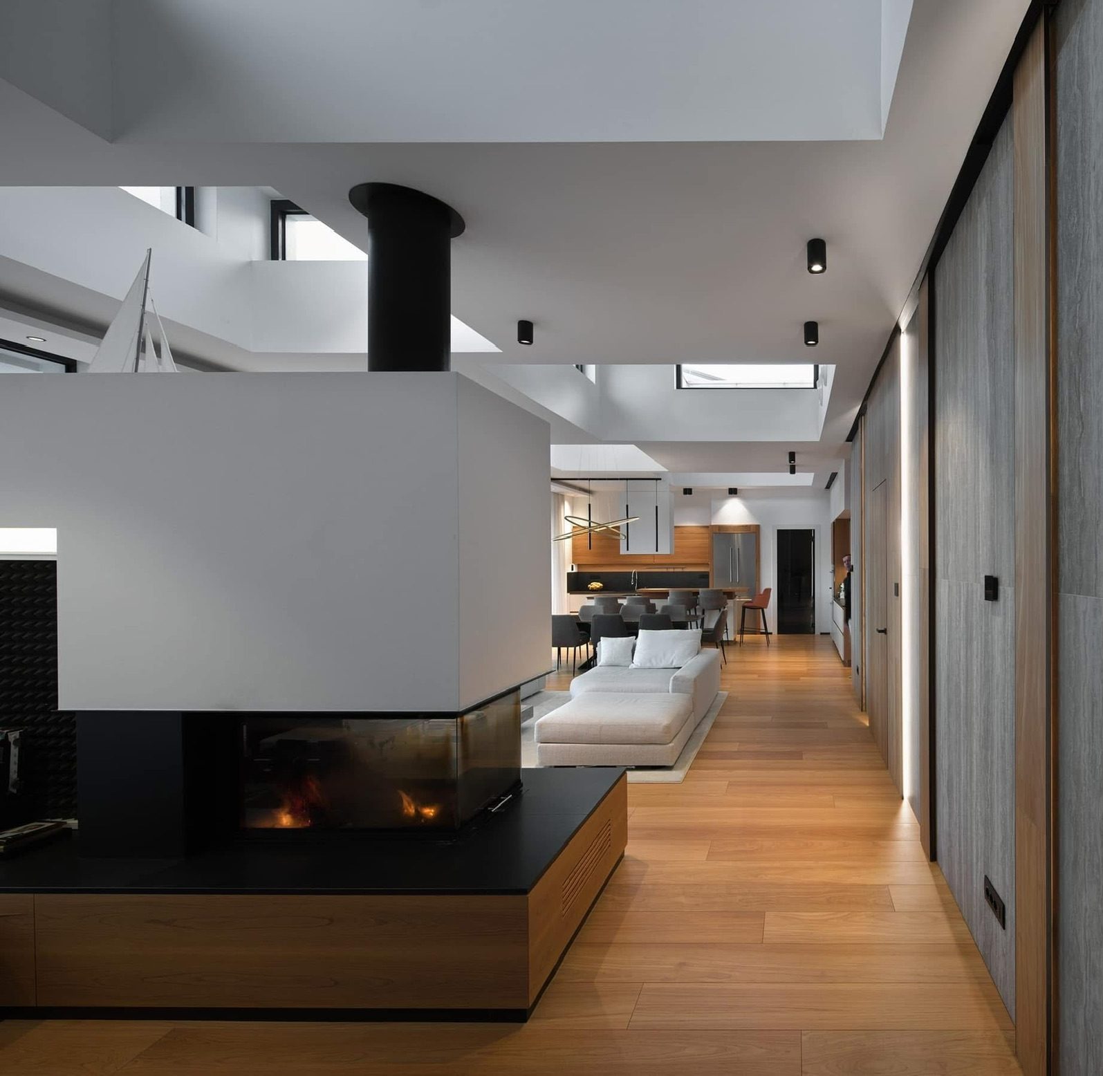 Moderan dnevni boravak otvorenog tipa s drvenim podovima, središnjim bijelim kaminom, sivom kutnom sofom, blagovaonicom i kuhinjom u pozadini, sve osvijetljeno prirodnom i ugradbenom rasvjetom. Elko – Instalacija Pametnih Kuća i Smart Home Sistema Moderan dnevni boravak otvorenog tipa s drvenim podovima, središnjim bijelim kaminom, sivom kutnom sofom, blagovaonicom i kuhinjom u pozadini, sve osvijetljeno prirodnom i ugradbenom rasvjetom. Elko - Instalacija Pametnih Kuća i Smart Home Sistema