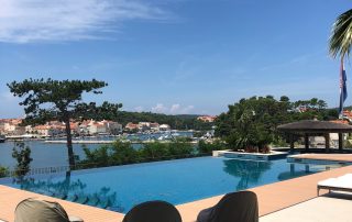 Infinity bazen s tri natkrivene ležaljke na popločanoj terasi, s pogledom na vodu, drveće i udaljeni grad pod plavim nebom. Elko - Instalacija Pametnih Kuća i Smart Home Sistema