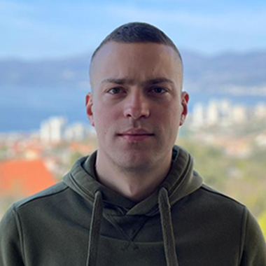 Muškarac s kratkom kosom u zelenoj majici s kapuljačom stoji vani s mutnim gradskim pejzažem i planinama u pozadini. Elko – Instalacija Pametnih Kuća i Smart Home Sistema Muškarac s kratkom kosom u zelenoj majici s kapuljačom stoji vani s mutnim gradskim pejzažem i planinama u pozadini. Elko - Instalacija Pametnih Kuća i Smart Home Sistema